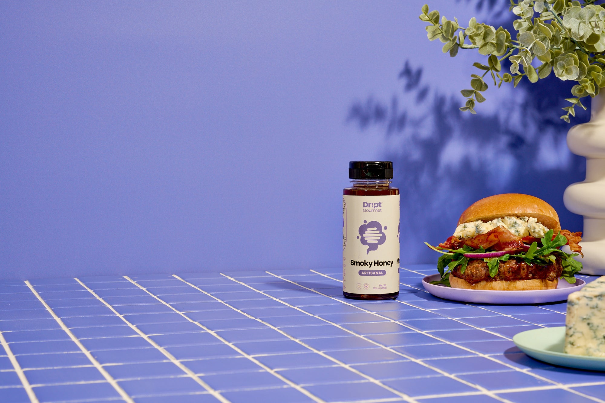 Dript Gourmet Smoky Honey with gourmet sandwich on a light blue tiled tables. Small-batch Canadian honey infused with smoky flavour.