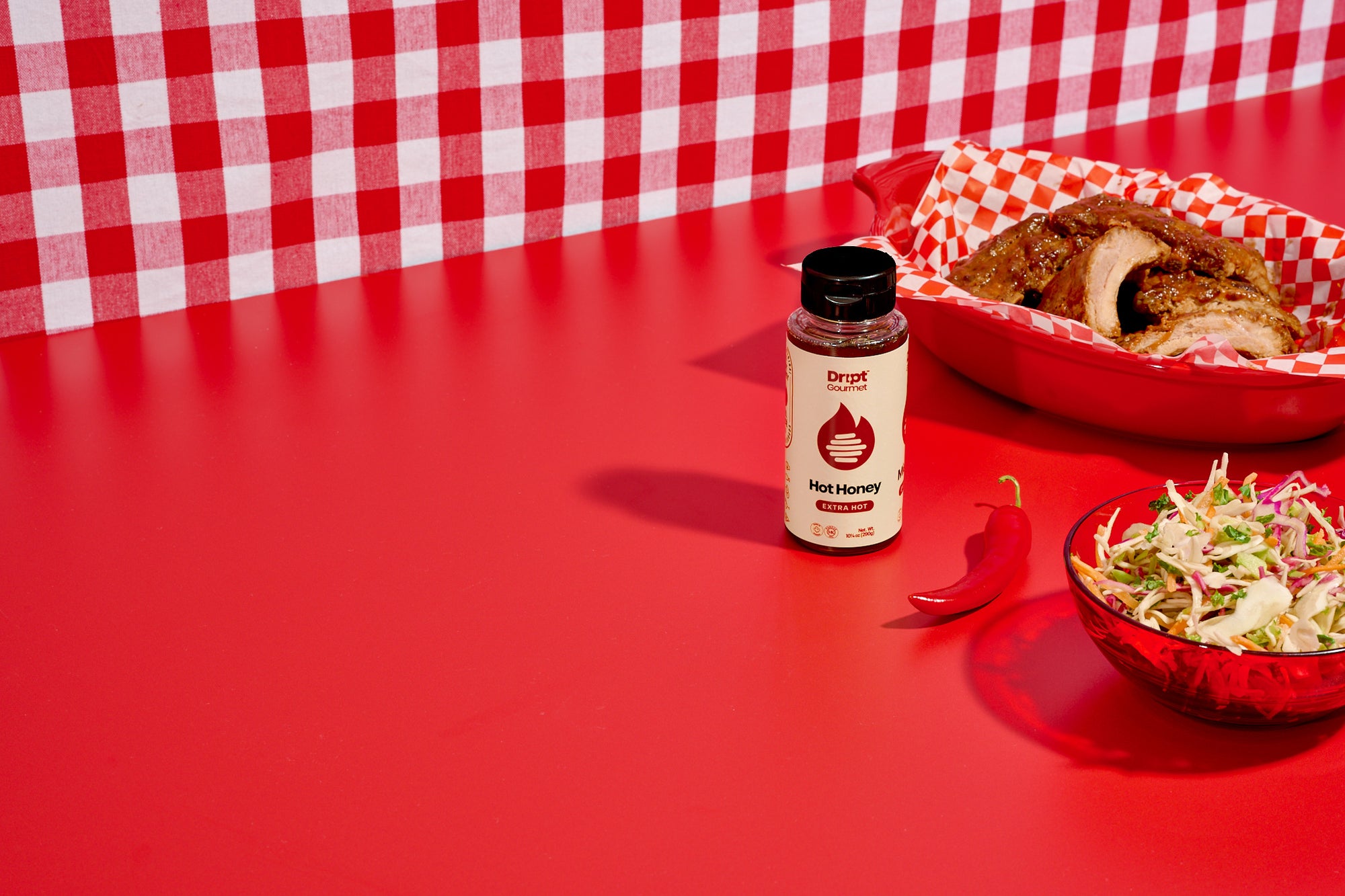 Côtes levées de miel extra piquantes Dript Gourmet sur un plateau rouge avec salade de chou, piment fort et une bouteille de miel extra piquant sur une table rouge.