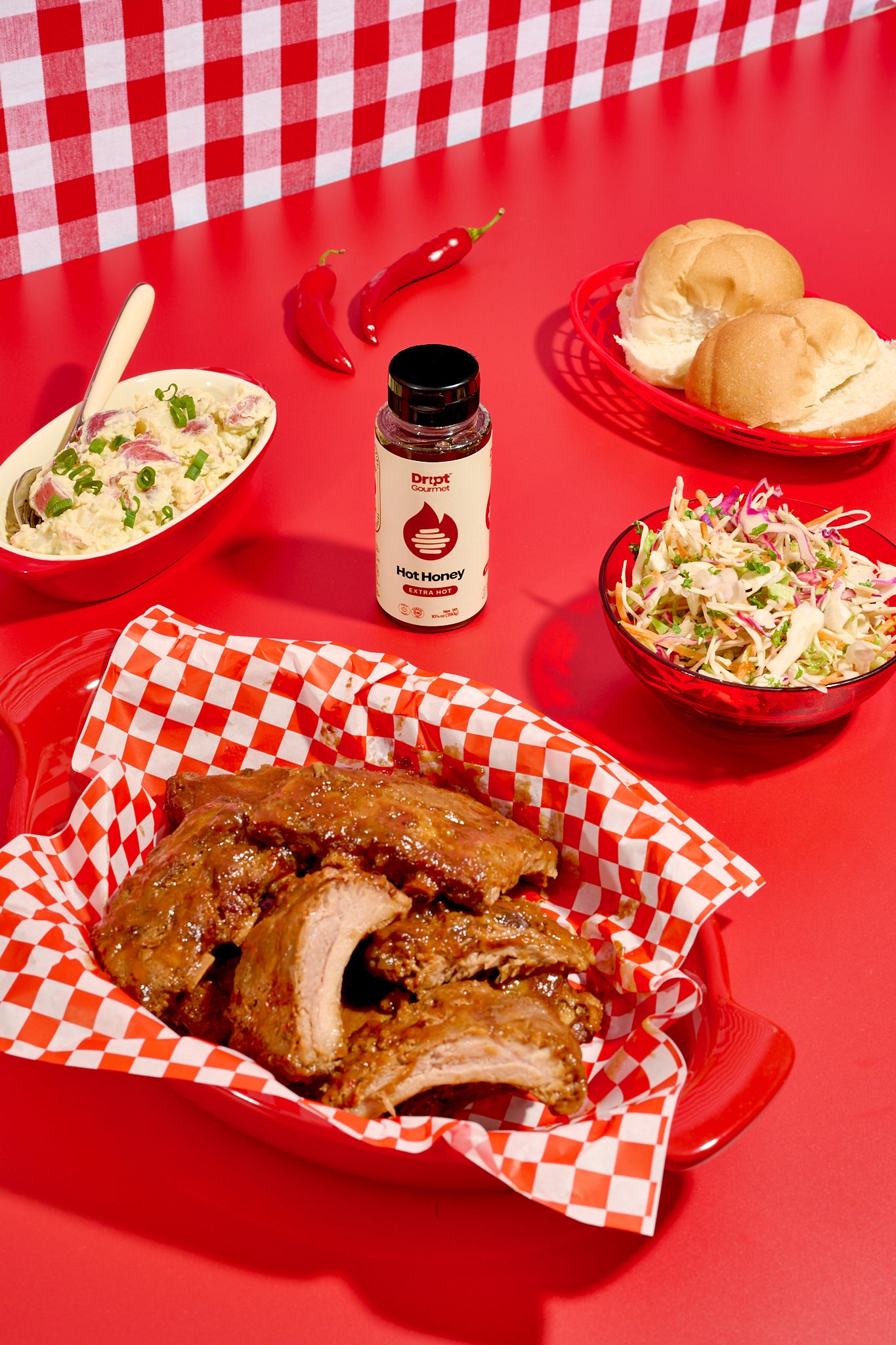 Dript Gourmet extra hot honey ribs on a red tray with coleslaw, buns, and a bottle of Extra Hot Honey on a red table.