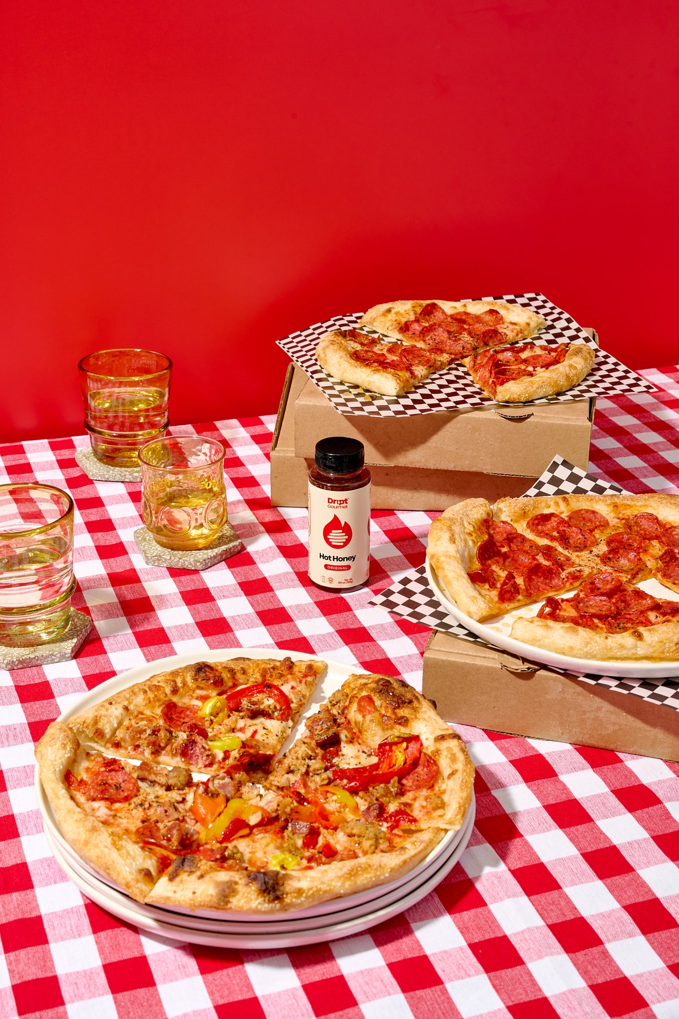 Dript Gourmet Original Hot Honey with 3 pizzas on a red and white diner tablecloth. Small-batch Canadian condiment adding bold sweet-heat flavour to pizza.