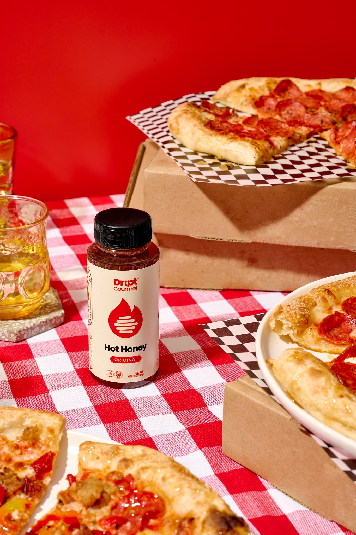 Dript Gourmet Original Hot Honey with 3 pizzas and pizza boxes on a red and white tablecloth. Small-batch Canadian condiment adding bold sweet-heat flavour to pizza.