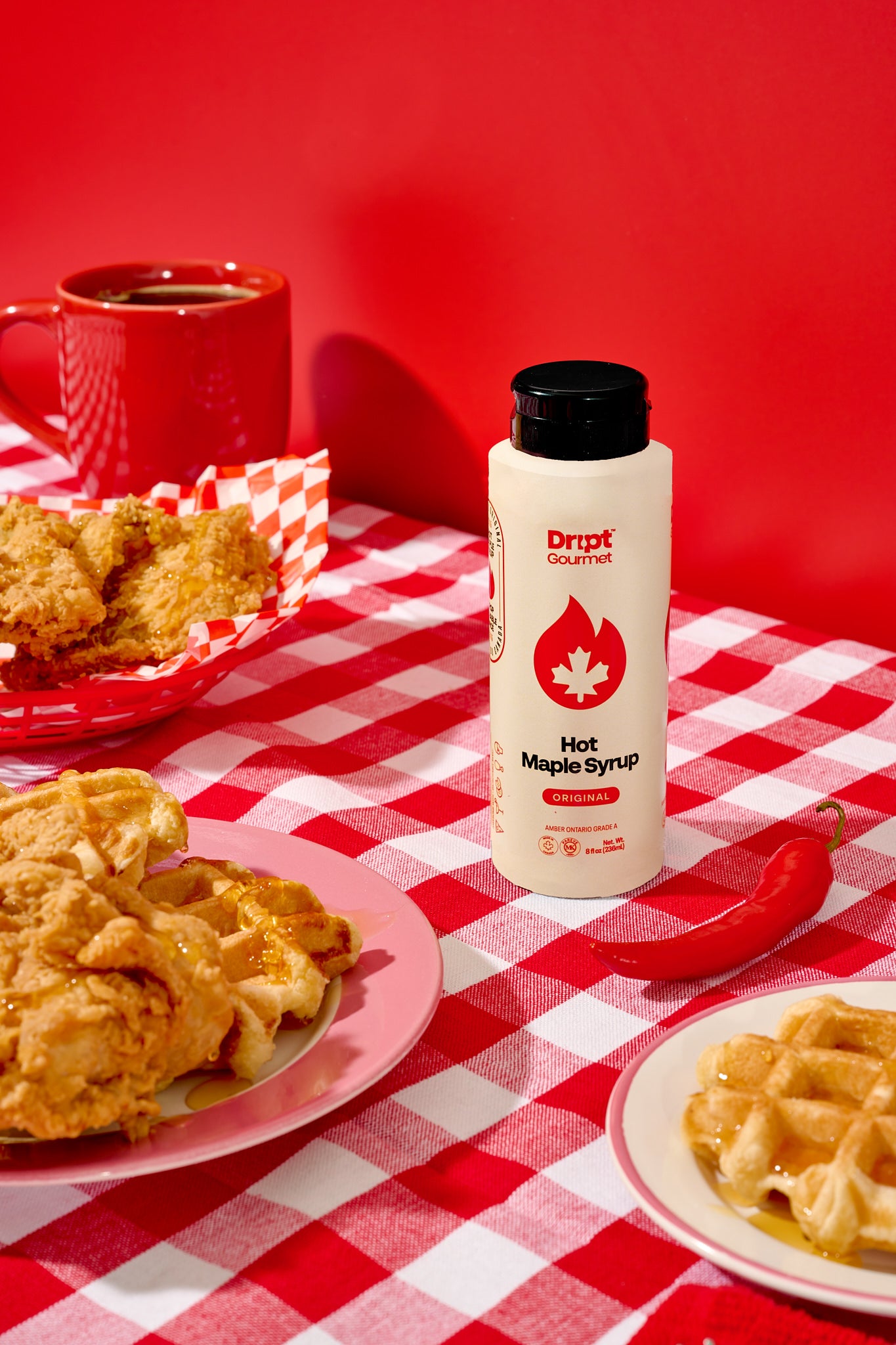 Fried chicken and waffles on a red and white checkered tablecloth with a bottle of Dript Gourmet Original Hot Maple Syrup.