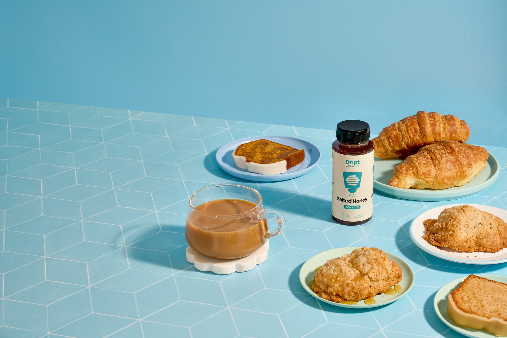 Dript Gourmet Salted Honey with croissants, coffee, and pastries on a light blue table. Small-batch Canadian honey infused with sea salt for sweet and savoury flavour.