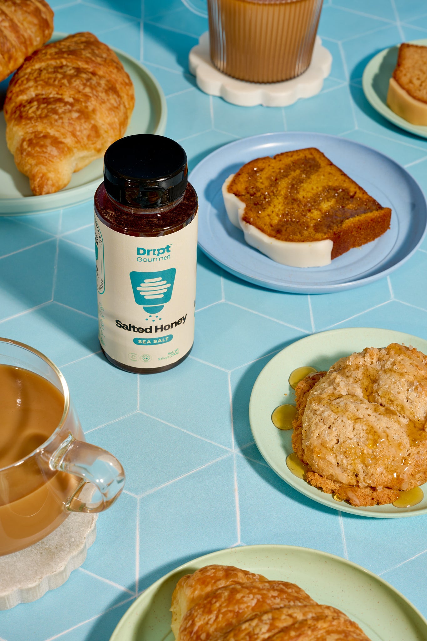 Dript Gourmet Salted Honey with croissants, coffee, and pastries on a light blue table. Small-batch Canadian honey infused with sea salt for sweet and savoury flavour.