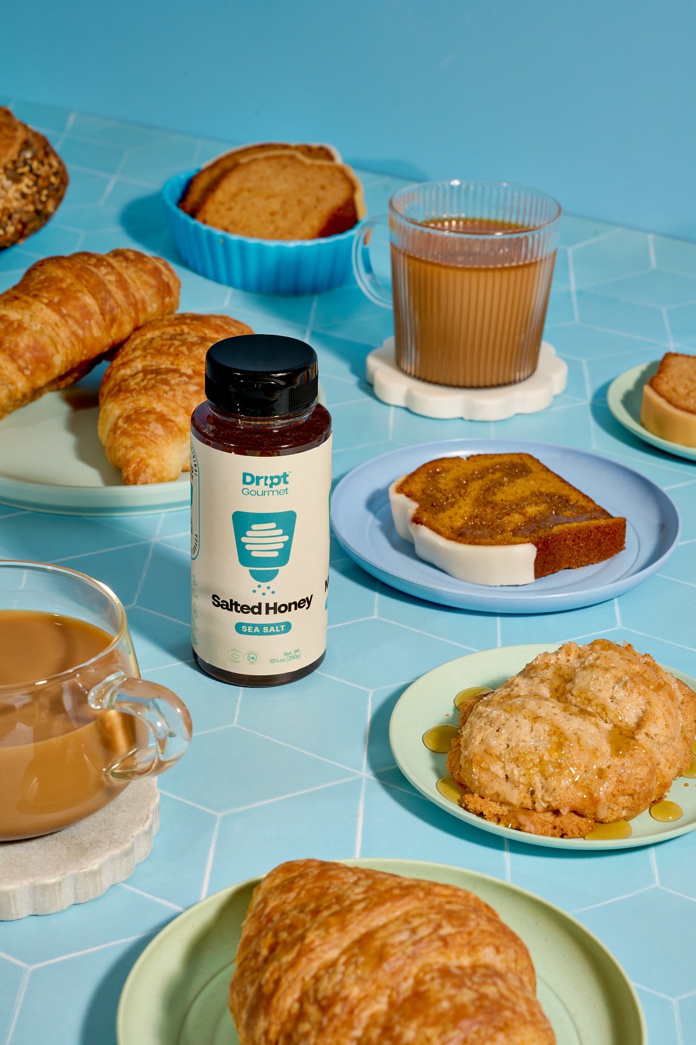 Dript Gourmet Salted Honey with croissants, coffee, and pastries on a light blue table. Small-batch Canadian honey infused with sea salt for sweet and savoury flavour.