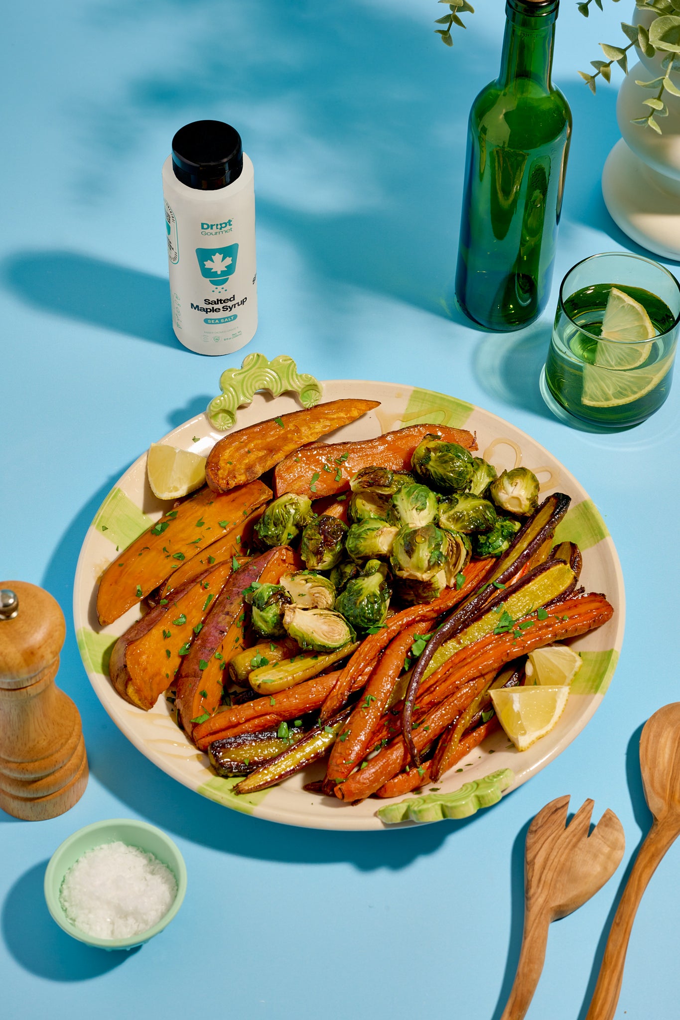 Dript Gourmet Salted Maple Syrup bottle with roasted carrots, sweet potatoes, and Brussels sprouts on a light blue table. Artisanal Canadian maple syrup infused with sea salt.